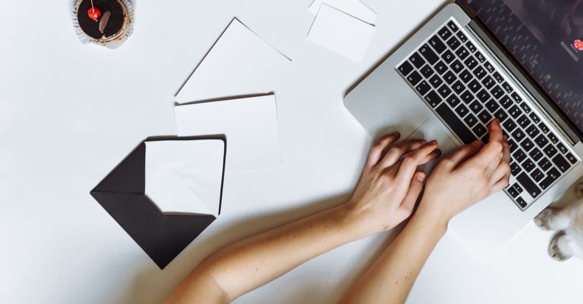 Top view of a person typing on a laptop with envelopes and a cupcake on a white desk.
